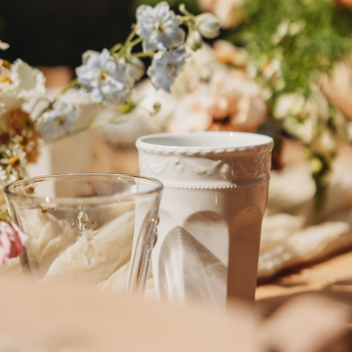 A glass and a white cup surrounded by flowers on a table.