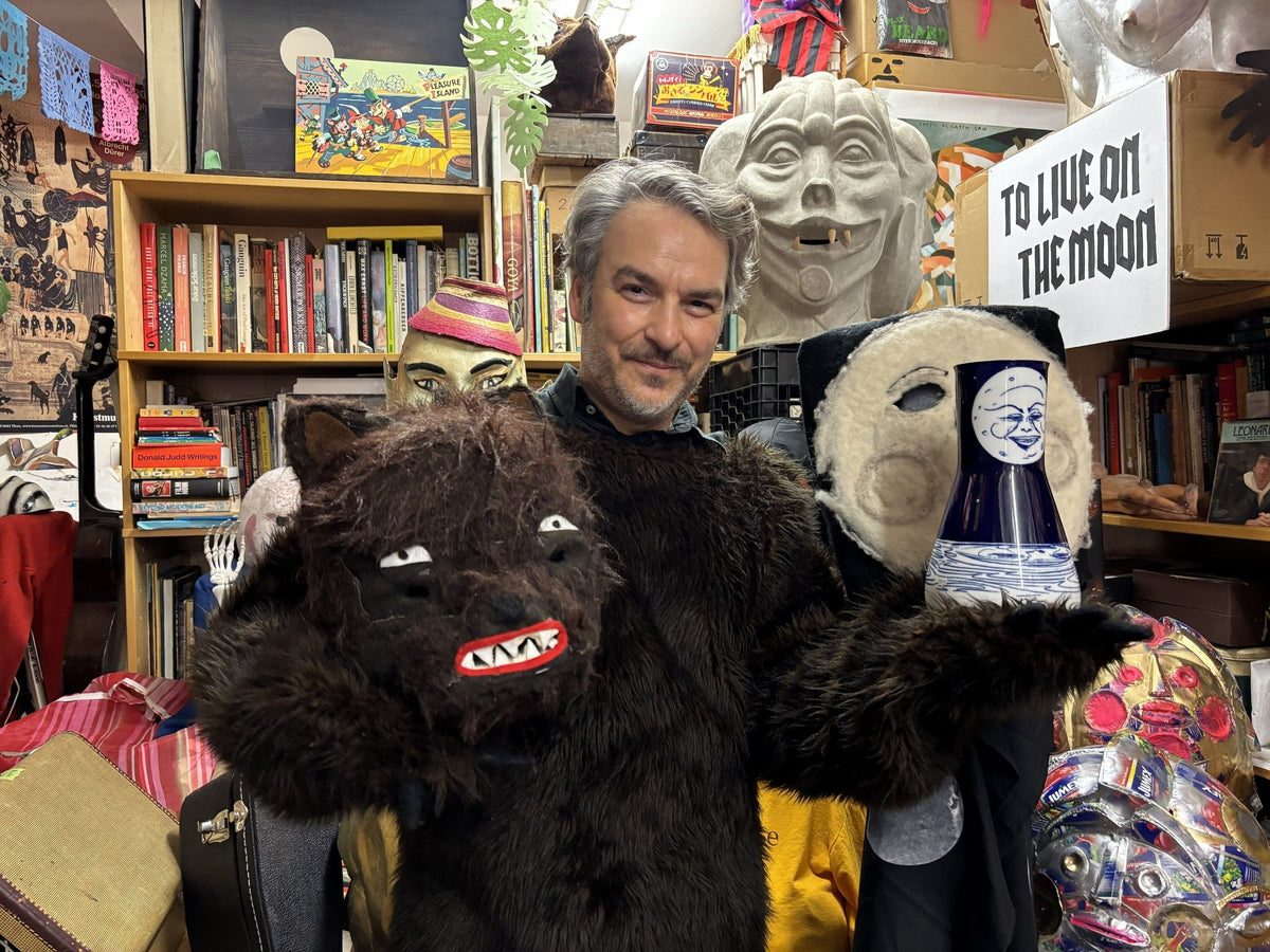 Person in a bear costume surrounded by various masks and bookshelves.