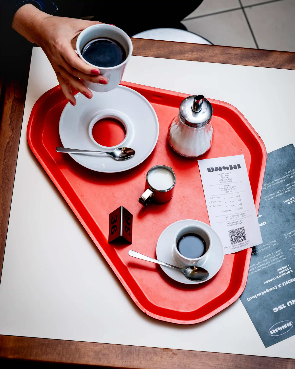 Eine Hand hält eine Flugzeugtasse auf einem roten Tablett mit Kaffee und Dessert.